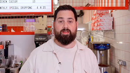 A picture of Rich Myers standing in his shop, behind a counter. There is a coffee machine and menu in the background.