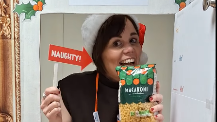 A woman with short, dark hair wearing a Santa hat smiling into the camera. She is holding an arrow with 'naughty' written on it pointing to a bag of macaroni in her other hand. She is standing behind a golden frame with holly in the top corners.
