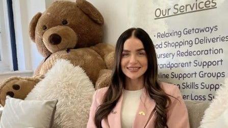 Aimee Green has dark hair and is smiling at the camera. She has a pink jacket and there is a bear behind her. There are some cushions near her.
