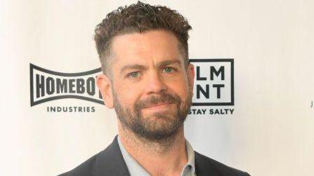 Jack Osbourne, who has dark hair cropped short and a beard, standing in front of a hoarding bearing various company logos. He is wearing a grey suit jacket and pale grey shirt and smiling.