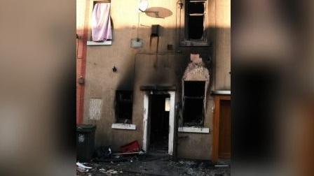 The charred and blackened front of the terraced house can be seen, with all of the windows and the door missing and blackened fire damaged materials on the pavement in front of the property.