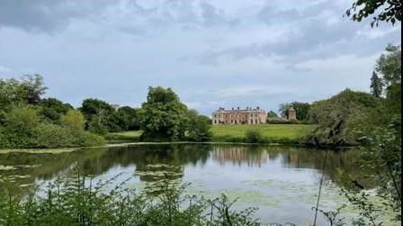 A large, stately home nestled on a grassy knoll is framed by trees with a lake in the foreground 