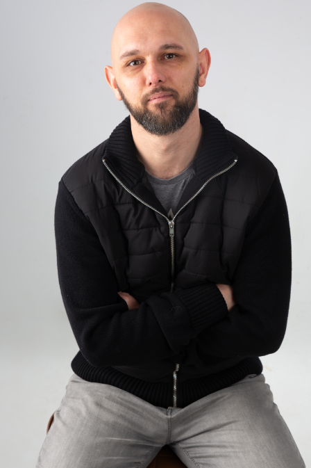 Man with bald head and a beard kneeling on a stool. He's wearing a black zip up top. 