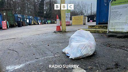 A white bag of rubbish lies abandoned outside a recycling centre. Blue skips can be seen in the background.