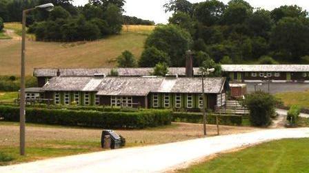 Rows of dormitary style buildings with slate rooves and green window frames sit in a green space in front of a gentle slope lined with trees.