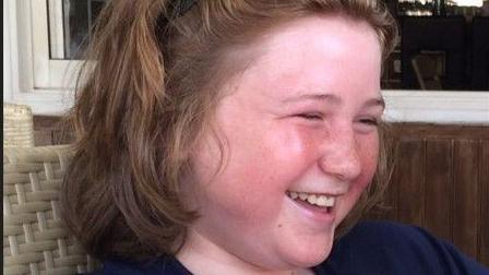 Ellis Cox as a child, with long brown hair, sitting in a wicker style chair and laughing