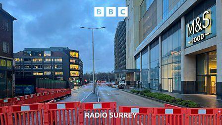 A line of red plastic barriers sit across a dual carriageway road. It's early morning and buildings line each side of the road. To the right the entrance to M&S food is clearly lit.