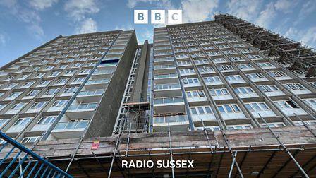 A block of flats rises into the sky. The flats look rundown and shabby and there is scaffolding around the base.