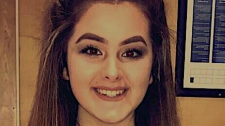 Emily smiles at the camera. She has dark eyeliner on and has thick long brown hair.