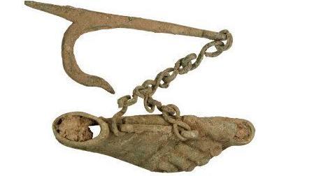 A rusted copper alloy carving of a foot, toenails included, attached to a diagonal chain hanging from a slim bar with a curved fork at the end