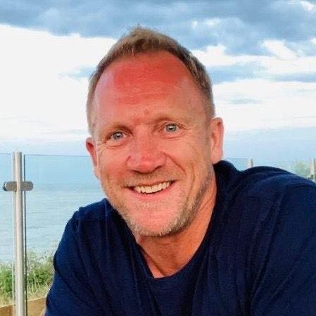 A man with sandy blond hair and a beard in a blue jumper smiles into the camera with the sea in the background