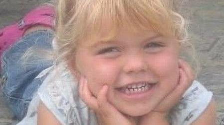 Elana is in the middle looking towards the camera smiling. She is laying on a concrete floor with her hands on her chin. She has blonde hair and a fringe.