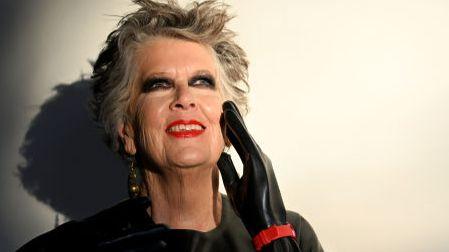 Prue Leith poses for a portrait photograph against a stark white background. Her black latex outfit contrasts with the background - with a flash of red detail to her wrist.