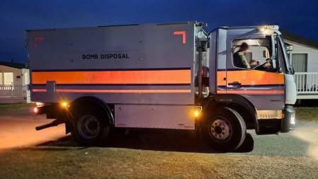 A large van with 'bomb disposal' written on the side. It is stopped outside what appear to be beach/holiday homes. The sky is a dark blue and it appears to be the evening. The van has its headlights on casting an orange glow onto the ground.