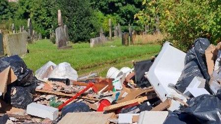 The image shows a large pile of dumped rubbish, scattered across the grass of a graveyard. The waste includes black bin bags, broken pieces of wood, plasterboard, household fixtures, cardboard, and various construction debris.
In the background, gravestones are visible.