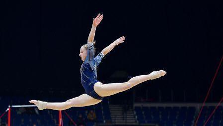 Olivia, performs a split leap on the floor exercise mat, with her arms extended gracefully. Gymnastics equipment, including uneven bars, is visible in the background, along with spectators and judges in a large arena setting.