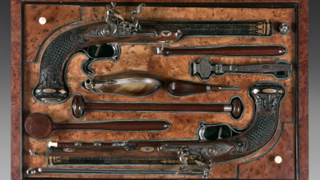 Two pistols lie in a wooden box alongside various accessories