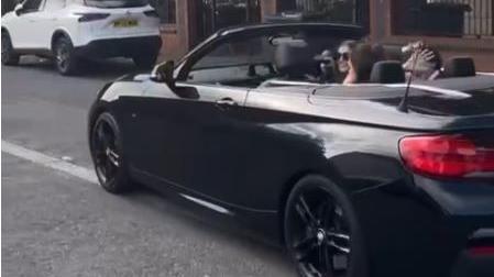 Screen grab of Hannah Taylor, wearing sunglasses, smiling as she drives a teenage girl and boy who are in the rear seats of her open-top convertible
