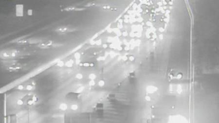 A black and white CCTV still of the overturned car. It has a red square superimposed around it and is lying on one of the road's two lanes. Emergency workers in hi-vis clothing are nearby, as are several of their vehicles.