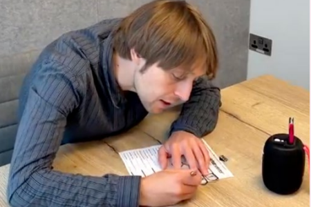 A man is filling in a ballot paper using a tactile voting aid, which is laid on top of the ballot paper, and an audio voting device which is placed next to him on a table. He has short, wavy brown hair and is wearing a blue striped shirt.