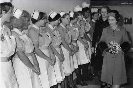 Queen Elizabeth II holds a bouquet of flowers as she walks past a line of student nurses, all wearing matching nurse uniforms and hats. The photo is in black and white.