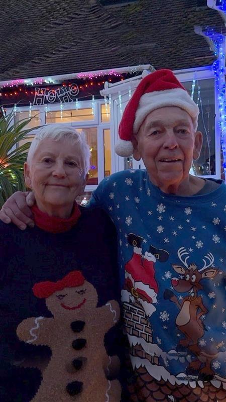An older couple wearing Christmas jumpers stand in front of their Christmas light covered bungalow.