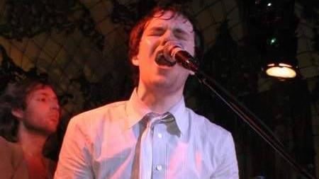 Brandon Flowers passionately singing into a microphone. He is wearing a white shirt and white tie, and stage lights illuminate the right side of his face with a pink hue. A bandmate looks on in the background.