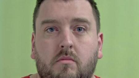 Police mugshot of Liam Bradley, who has close-cropped brown hair and a beard, photographed against a green background. 