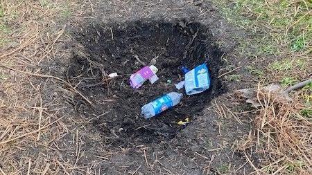 A plastic bottle and food wrappers left in a hole in the ground that has been freshly dug.