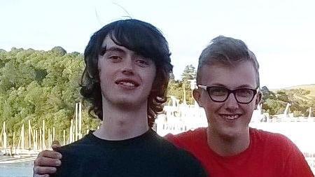 Two teenage boys next to each other in an outdoor setting smiling at the camera. Tom, left, has long hair and is wearing a black shirt. His brother wears a red shirt.