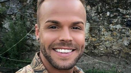 A close up photo of a man with stubble and a brown shirt