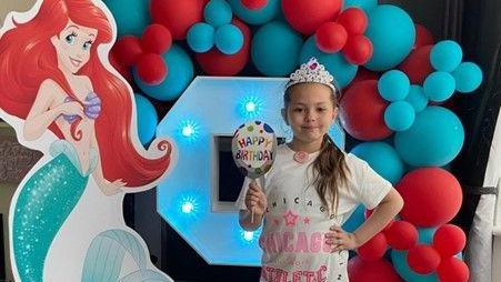 Olivia stands in front of an LED number nine figure which is surrounded  by red and blue balloons and a cardboard cut out of Princess Ariel. Olivia has her hand on her hip and is wearing a white t-shirt with pink wording and a crown.