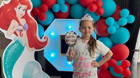 Olivia wears a tiara and a white-and-pink t-shirt. She smiles as she holds a Happy Birthday sign in front of an arch of red and blue balloons, next to a large cutout of Ariel from the animated Disney film The Little Mermaid.