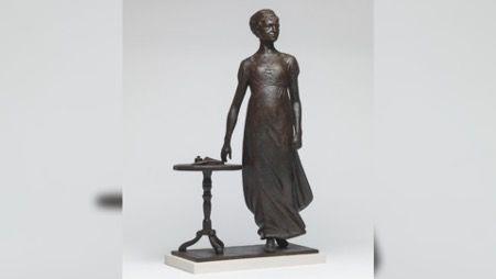 A statue of a woman in a long dress standing to the right of a small round table which has paper and an inkwell on it