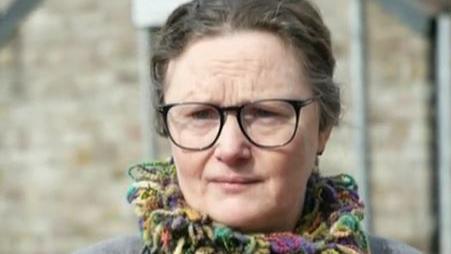 A headshot of Katie Sullivan. She is wearing a multi-coloured scarf and glasses and is unsmiling.