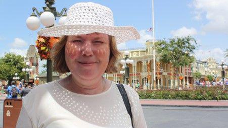 A woman wearing a white sun hat and T-shirt stands outside on a sunny day. A building and trees can be seen behind her. People are crossing the road.