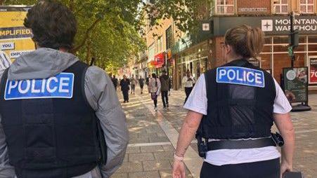 Male police officer wearing a black vest with a blue police sign and a grey hoodie and a woman officer wearing the same police vest and a white T-shirt walking along Bridge Street in Peterborough 
