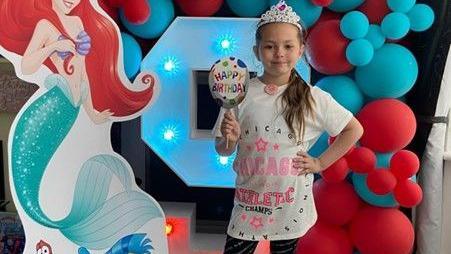 Olivia stands in front of an LED number nine figure which is surrounded  by red and blue balloons and a cardboard cut out of Princess Ariel. Olivia has her hand on her hip and is wearing a white t-shirt with pink wording and a crown.