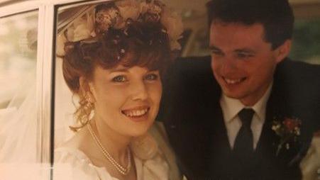 A photo of a bride and groom in a car on their wedding day. The woman has wavy red hair tied up and is wearing a pearl necklace, floral headpiece and a white wedding dress and is smiling into the camera. The man has short brown hair and is wearing a dark suit and tie with a white shirt and floral pin. He is looking wistfully at the bride.