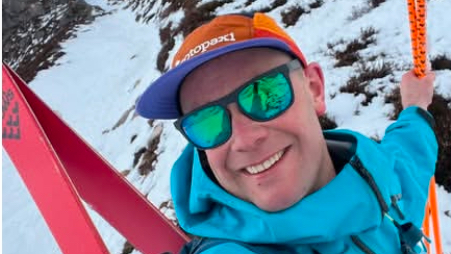 Selfie of Sam Burns, who is outside on a snowy slope. He is smiling and wearing an orange and purple cap, reflective sun glasses and a light blue jacket.