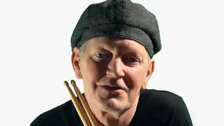 John Bennetts holding drumsticks and wearing a black t-shirt and grey hat, looking at the camera