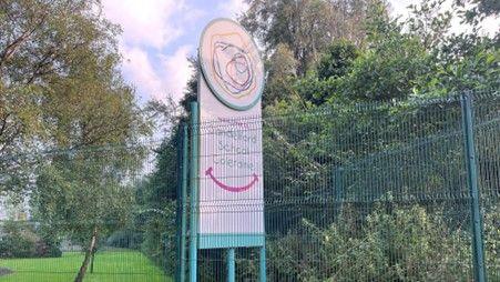 Sandelford school sign. The sign is white with green writing and is behind a green fence. There are trees in the background.