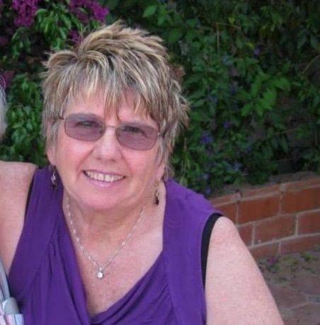 Lin McNally, with short blonde hair, sun glasses, a silver necklace and purple sleeveless top, smiles at the camera.