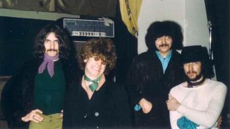 One man with curly hair, green pants and a green sweatshirt, stands next to a smiling Ozzy Osbourne who is wearing sunglasses and a green neck-tie, two other men are to his right. There's an amp and stage behind them.