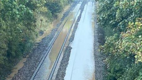 Flood water is sitting on top of railway lines travelling in both directions. There is also flood water next to a treeline on the left of the railway tracks. There are trees on either side of the railway.