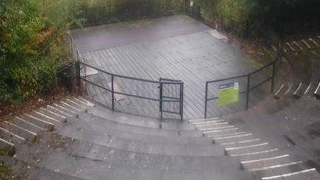 An amphitheatre in the rain. There is black metal fencing around the stage area with several layers of seating behind it, with steps down the centre.