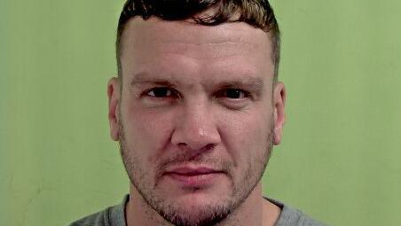 Donovan Kitching, who has dark brown short hair and stubble and is wearing a grey sweatshirt, in a police mugshot against a green background.