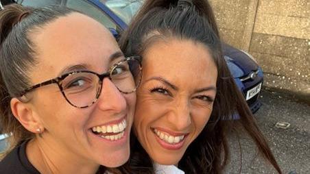 Amie Gray and her wife Sian Gray - both have brown hair that is tied back and they are smiling at the camera. Mrs Gray is wearing glasses