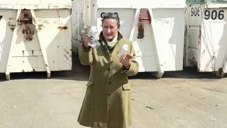 Councillor Elissa Swinglehurst wearing a green tweed coat holds up rubbish in both hands while standing in front of white recycling bins.