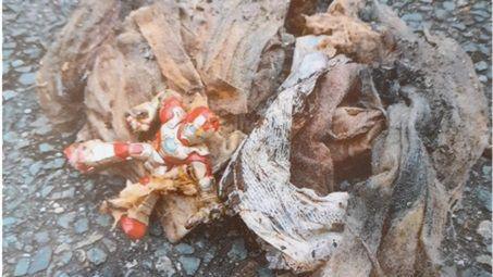 An Ironman plastic figure is lying on tarmac with a bundle of what looks like twisted and dirty fabric wipes.